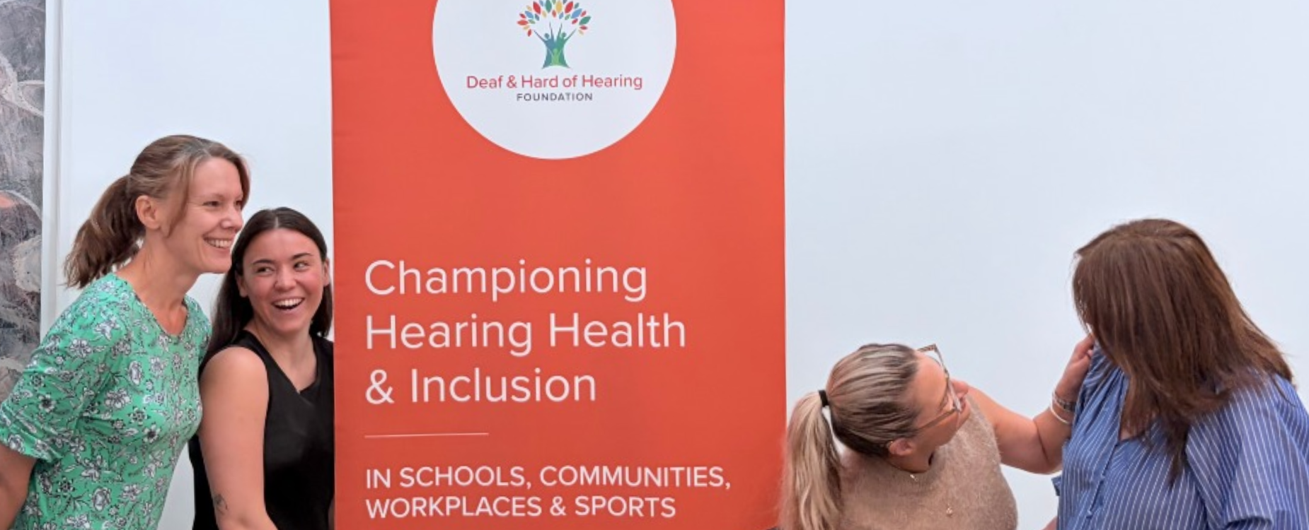 Five staff members standing beside a Deaf and Hard of Hearing Foundation banner and being playful.